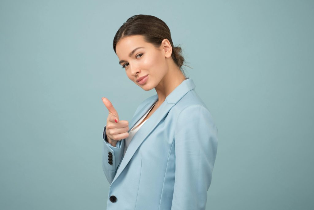 Young brunette woman wearing a light blue blazer standing infront of a wall painted the same colour. She is winking and pointing at the camera. She looks confident.