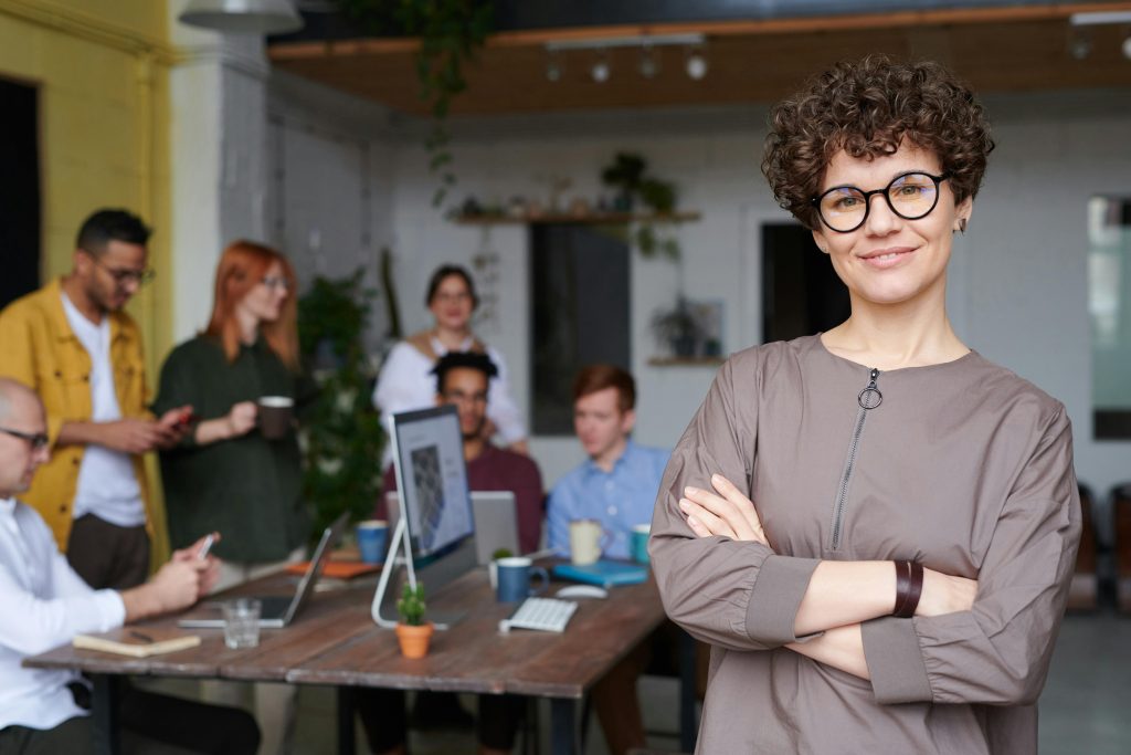 Smiling woman with brown hair and glasses stands with her arms folded, looking confident. The background is out of focus but shows a team of people collaborating around a desk.