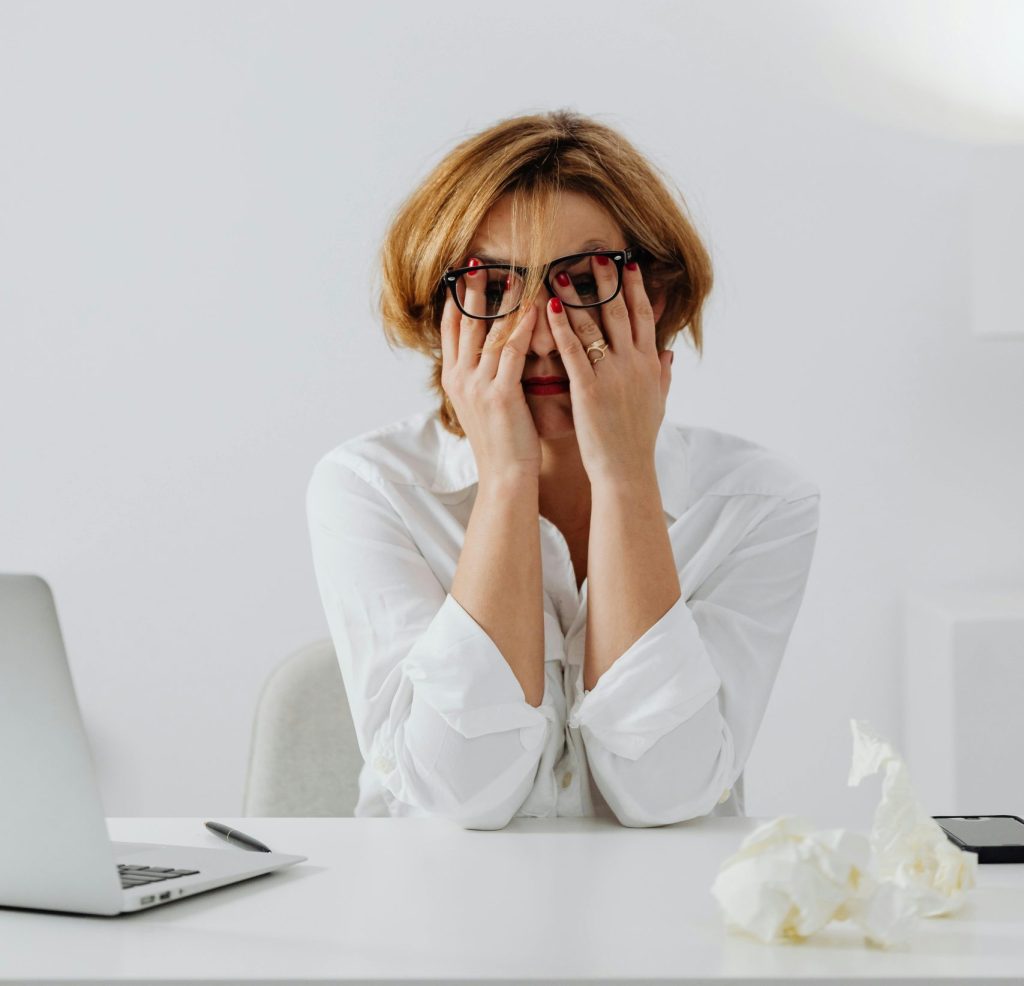 A blonde woman sits with her hands covering her face. Her glasses sit ontop of her hands. She looks stressed. On the desk by her is an open laptop and a crumpled piece of paper.
