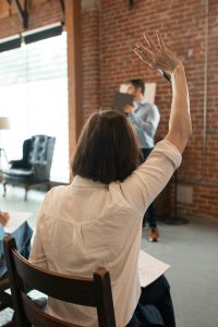 Woman sat with her back to us, facing a stage. She is raising her hand to answer a question. The presenter is in front of her.
