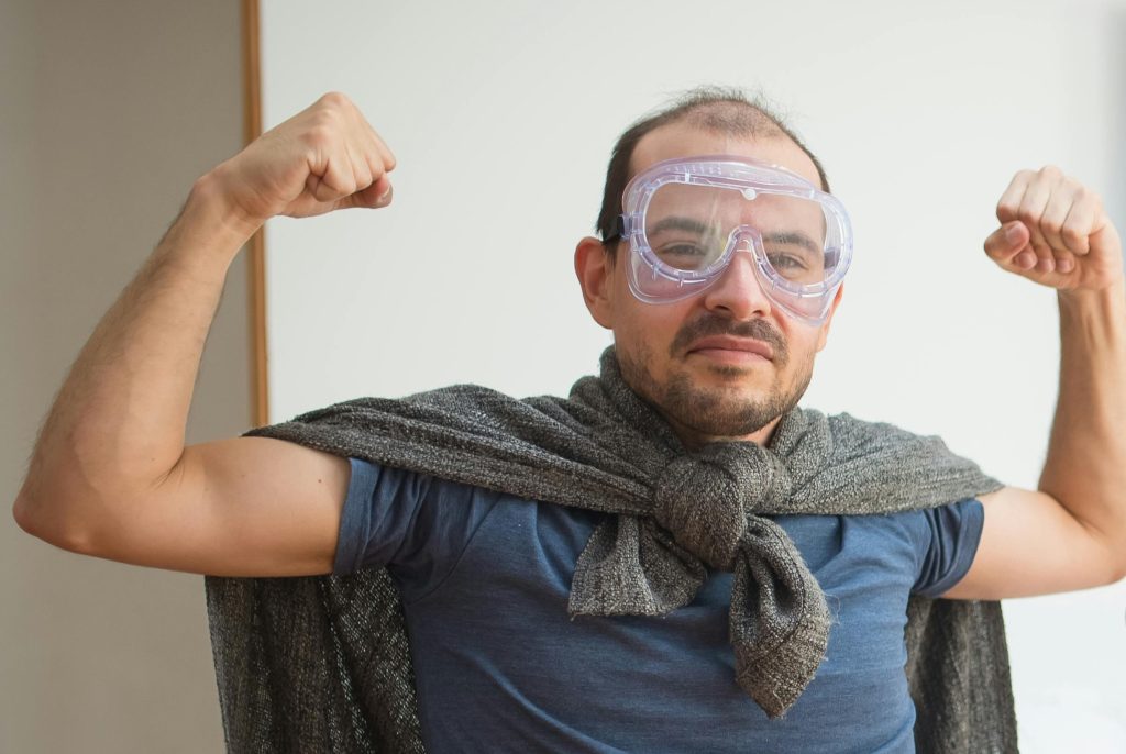 Middle-aged white man playing superhero dress-up with a jumper as a cape and DIY goggles as a mask. He is flexing his biceps and about to laugh.